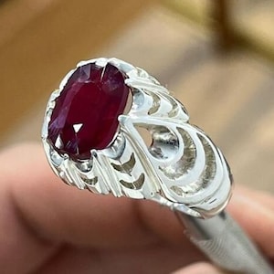 Peut inclure: Bague en argent ornée d'une pierre précieuse rouge foncé de forme ovale. La bague présente un motif ajouré ornemental sur le corps, avec une finition polie. La pierre précieuse est le point central, mettant en valeur sa couleur riche et sa clarté.
