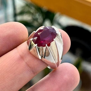 Könnte beinhalten: Ein silberner Ring mit einem großen, ovalen Rubin-Edelstein. Der Ring hat ein detailliertes, geometrisches Design.