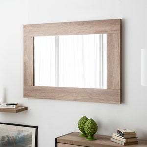 Könnte beinhalten: Ein rechteckiger Spiegel mit einem Holzrahmen in hellbrauner Holzoptik. Der Spiegel hängt an einer weißen Wand über einem Holzregal mit einer Vase und einem Buch. Eine Tischlampe mit einem weißen Lampenschirm befindet sich auf der rechten Seite des Bildes.