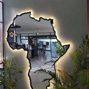 May include: A wall-mounted mirror shaped like the continent of Africa, featuring a black frame and a warm white LED backlight that illuminates the perimeter. The reflective surface shows a clear view of an indoor space with plants and lighting.