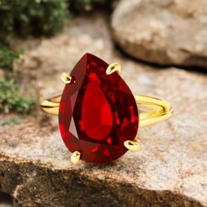 May include: A gold ring featuring a large, pear-shaped, faceted red gemstone. The ring's band is a polished gold, and the gemstone is held in place by four gold prongs.