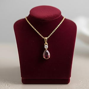 May include: A gold-colored necklace with a teardrop-shaped, deep red gemstone pendant. A small, clear gemstone sits above the red stone. The necklace is displayed on a burgundy velvet jewelry stand.