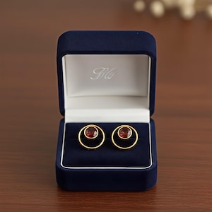 May include: Gold-coloured earrings with a red gemstone centre, presented in an open navy blue jewellery box. The earrings feature a circular design. The box has a white interior and a logo.