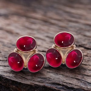 May include: A pair of gold-coloured stud earrings, each with three oval, deep red gemstones. The earrings are set against a rustic wooden background, highlighting the jewellery's design and colour.