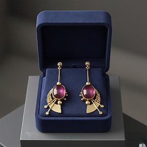May include: Gold-coloured earrings with large, oval, magenta stones, presented in a dark blue velvet jewellery box. The earrings feature a distinctive design with small gold beads and geometric shapes, suitable for special occasions.