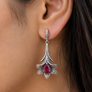 May include: A silver-toned drop earring featuring an ornate, floral-inspired metal design with intricate scrollwork detailing. At the center of the flower-shaped base sits a single, teardrop-shaped, deep red gemstone held by a metal prong setting.