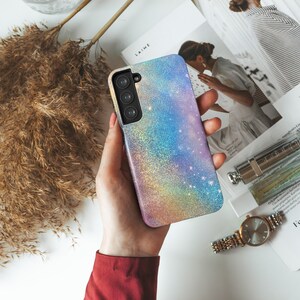 Może przedstawiać: Etui na telefon z błyszczącym wzorem tęczy. Etui ma przezroczyste plecy i czarny wycięcie na aparat.