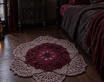 May include: A round, crocheted rug featuring a mandala-inspired pattern with concentric rings of deep burgundy, magenta, and light mauve yarn. The design includes intricate openwork stitches and a scalloped edge, creating a textured, floral-like shape.
