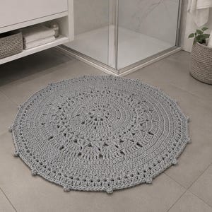 Könnte beinhalten: Ein runder, hellgrauer Häkelteppich mit einem detaillierten, sich wiederholenden geometrischen Muster, einer strukturierten, geknoteten Oberfläche und einem gewellten Rand, der flach auf einem Fliesenboden vor einem modernen Badezimmerschrank liegt.