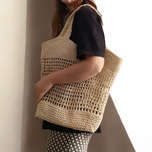 May include: A beige, open-weave tote bag with a rectangular shape and two shoulder straps. The bag is made of a woven material with a large, open pattern. The bag is being held by a person.