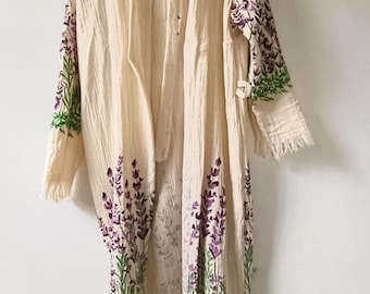 May include: A long, cream-coloured robe made of textured, crinkled fabric. The garment features long sleeves and a front opening, decorated with painted purple lavender sprigs and green stems along the lower hem and the outer sleeve cuffs.