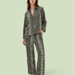 Puede incluir: Una camisa de manga larga con botones y pantalones anchos a juego con un estampado continuo de piel de serpiente en negro, gris y verde oliva. El conjunto se completa con zapatos planos de color marrón oscuro, creando un estilo coordinado.