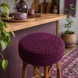 May include: A round stool featuring a thick, dark purple crocheted seat cushion atop a wooden base with four legs and a circular footrest. The seat is crafted from textured yarn in a dense, circular pattern, providing a firm, elevated surface.