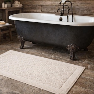 May include: A cream-coloured rectangular bath mat with a geometric pattern sits in front of a dark grey claw-foot bathtub. The bathroom has a rustic, spa-like aesthetic with wooden accents and a vintage tap.