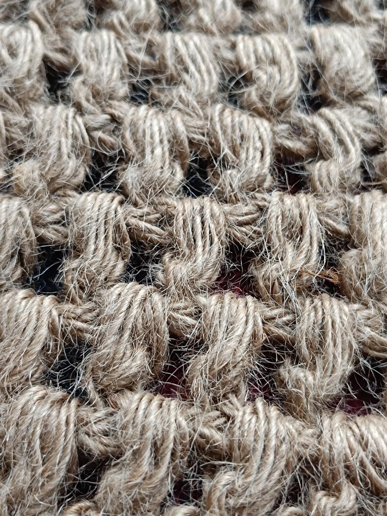 May include: Close-up of a woven jute rug with a natural brown color. The rug has a textured, rough surface and is made of tightly woven strands of jute.
