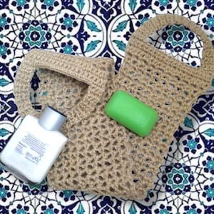 May include: A beige crocheted mesh bag with two handles, featuring a rectangular green bar of soap and a white plastic bottle with a silver cap placed on top of the open-weave fabric structure of the bag.