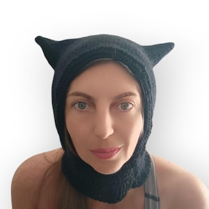 May include: A black knitted balaclava featuring two pointed cat-like ears on the top. The headgear is constructed from a textured yarn material and covers the head and neck area, framing the face with a snug fit and a rounded opening for visibility.