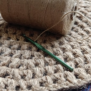 May include: A close-up of a crocheted rug made with natural jute twine. A ball of twine and a green crochet hook are lying on the rug.