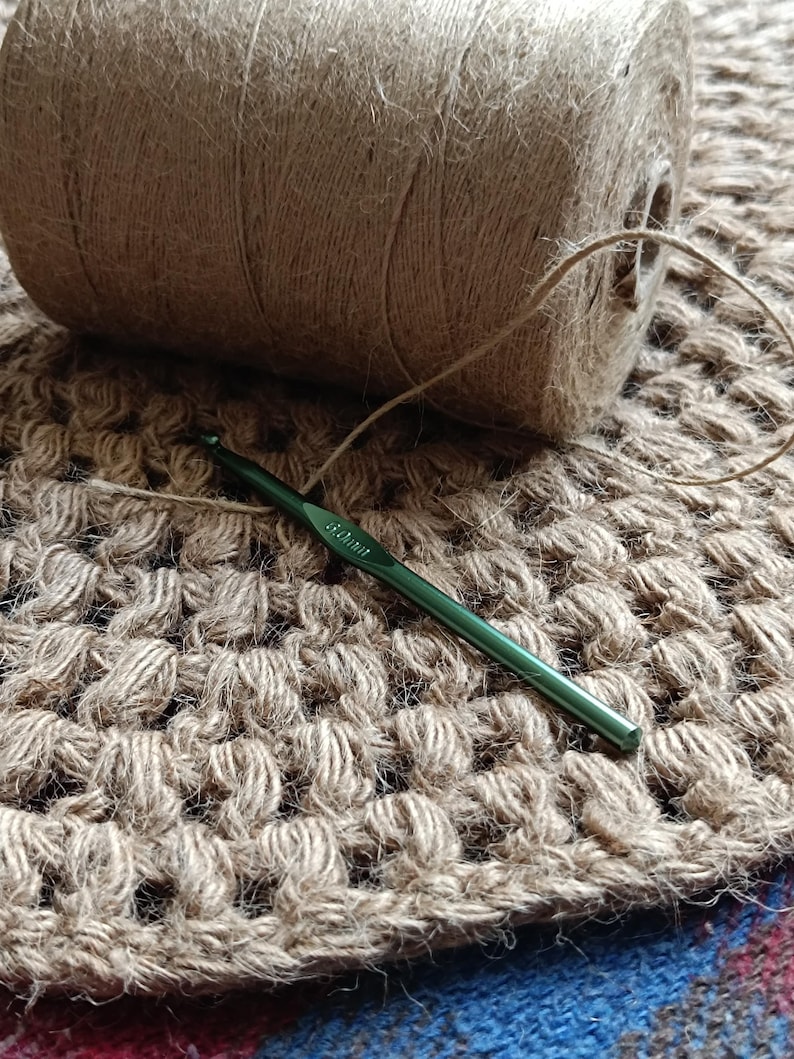 May include: A close-up of a crocheted rug made with natural jute twine. A ball of twine and a green crochet hook are lying on the rug.