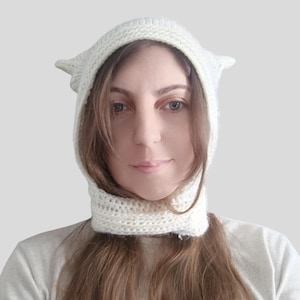 May include: A cream-colored, hand-knitted balaclava-style hood featuring two pointed, cat-ear-shaped protrusions at the top. The headpiece is crafted from a thick, textured yarn and fits snugly around the face, extending down to cover the neck area.