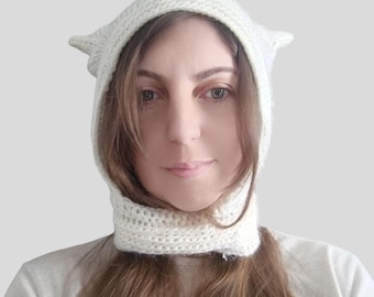 May include: A cream-coloured, hand-knitted balaclava-style hood featuring two pointed, cat-ear-shaped protrusions at the top. The headpiece is crafted from a thick, textured yarn and fits snugly around the face, extending down to cover the neck area.