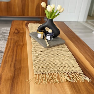 Pode incluir: Um caminho de mesa retangular de crochê bege com franjas nas extremidades, coberto por uma bandeja retangular cinza, um vaso preto abstrato contendo tulipas artificiais cor de creme e dois pequenos frascos de vidro com etiquetas.
