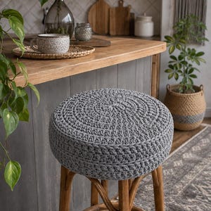 May include: A round, gray crocheted stool cover featuring a dense, textured stitch pattern. The cover is fitted onto a wooden stool with four legs and a circular support brace, providing a thick, cushioned seat surface with a uniform gray color.