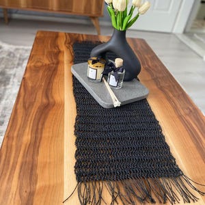Peut inclure: Un chemin de table noir au crochet avec des franges, surmonté d'un plateau rectangulaire en pierre grise. Le plateau contient un vase noir mat avec des tulipes crème, une bougie dans un pot en verre à couvercle doré et un diffuseur à roseaux.