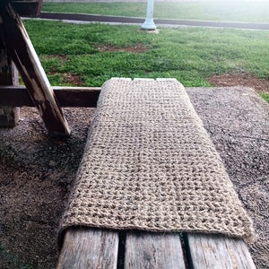 May include: A hand-crocheted bench cover in a natural, light brown color. The cover is made of a textured, open-weave material and is draped over a weathered wooden bench. The bench is outdoors, with a grassy area in the background.