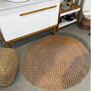 May include: A round, beige, crocheted rug with a textured, woven design. The rug is placed on a light brown floor in a bathroom setting.