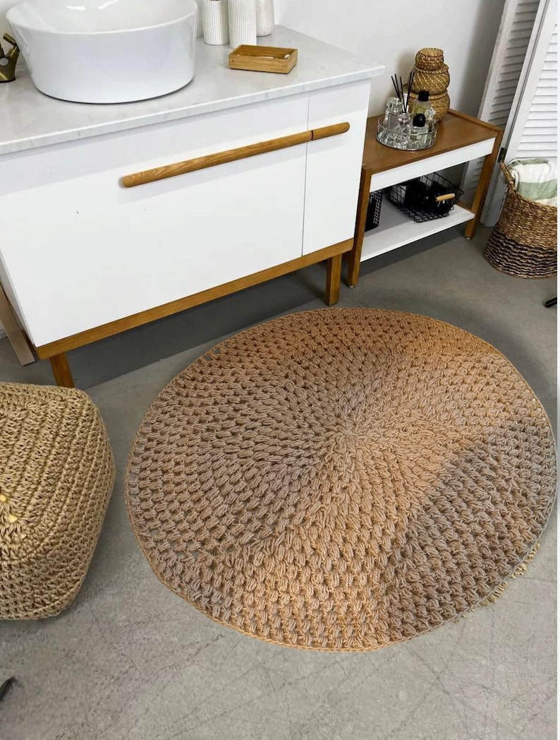 May include: A round, beige, crocheted rug with a textured, woven design. The rug is placed on a light brown floor in a bathroom setting.