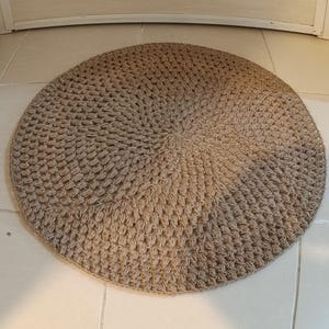 May include: A round, beige, crocheted rug with a textured surface. The rug is made of natural fibers and has a circular design.