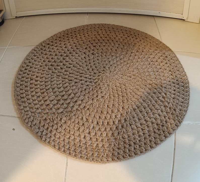 May include: A round, beige, crocheted rug with a textured surface. The rug is made of natural fibers and has a circular design.