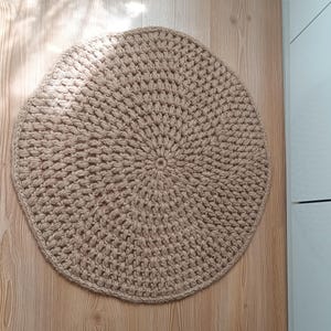 May include: A round, beige crocheted rug with a circular pattern. The rug is made of natural fibers and has a rustic, handmade look.