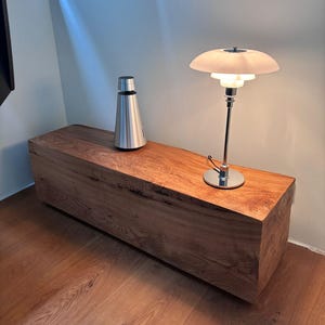Puede incluir: Una estantería de madera con un altavoz plateado y una lámpara cromada con una pantalla blanca. La estantería está hecha de madera natural con un acabado rústico.