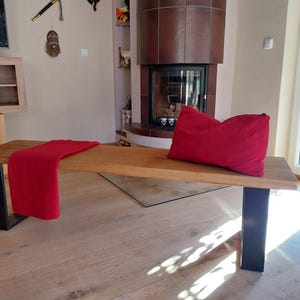 K&ouml;nnte beinhalten: Eine Holzbank mit schwarzen Metallbeinen. Die Bank ist aus Naturholz gefertigt und hat eine rote Wolldecke darauf. Die Bank steht in einem Wohnzimmer mit einem Kamin im Hintergrund.
