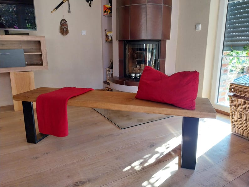 K&ouml;nnte beinhalten: Eine Holzbank mit schwarzen Metallbeinen. Die Bank ist aus Naturholz gefertigt und hat eine rote Wolldecke darauf. Die Bank steht in einem Wohnzimmer mit einem Kamin im Hintergrund.