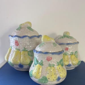 Può includere: Tre barattoli di ceramica con coperchi. I barattoli sono bianchi con un motivo a intreccio di cestino, pere gialle e accenti blu e verdi. I coperchi hanno una maniglia a forma di pera gialla.
