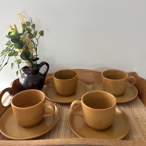 Può includere: Quattro tazze da caffè e piattini in ceramica maculata abbinati, in una calda tonalità terrosa. Le tazze hanno corpi arrotondati e piccole maniglie. Sono disposte su un vassoio di legno con una brocca marrone scuro e vegetazione sullo sfondo.