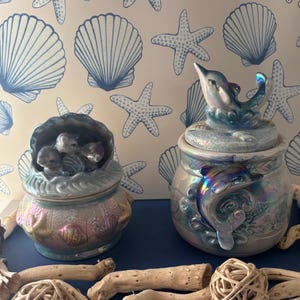 Peut inclure: Deux pots en céramique irisés avec des motifs de dauphins et de coquillages. L'un des pots présente des dauphins à l'intérieur d'une coquille, l'autre a un dauphin sur le dessus. Les pots sont sur une surface bleue avec du bois flotté et une boule tressée.
