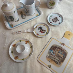 Könnte beinhalten: Eine Sammlung weißer Keramik-Tischutensilien mit Illustrationen von Hasen und Waldtieren. Enthält eine Tasse, ein Tablett, kleine Teller, einen Löffel, eine Zuckerdose und ein Schneidebrett mit gelbem Rand.
