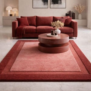May include: A living room scene featuring a red sofa with matching throw pillows and a blanket. A round, tiered coffee table sits on a large, two-tone red rug. The rug has a lighter center and a darker border. Two abstract art pieces hang on the wall.