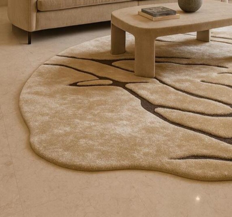 Pode incluir: Um tapete redondo bege com um design org&acirc;nico &uacute;nico. Padr&otilde;es castanhos escuros, semelhantes a veias, s&atilde;o tecidos no tapete macio de cor clara. O tapete est&aacute; colocado sobre um ch&atilde;o de azulejos claros, com um sof&aacute; e uma mesa de centro bege no fundo.
