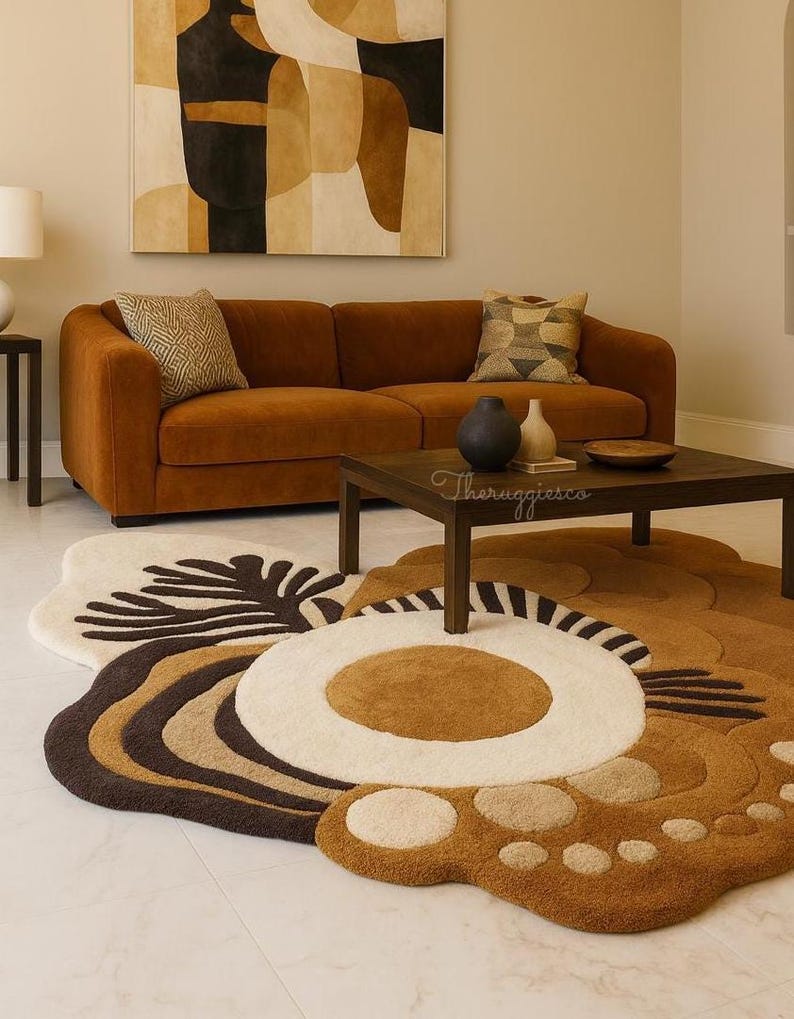 May include: A living room scene featuring a rust-colored sofa, a dark wood coffee table, and a large, floral-shaped rug in shades of brown, cream, and white. A painting with abstract shapes hangs on the wall above the sofa.