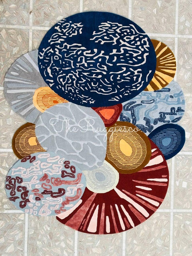 May include: A collection of circular rugs in various sizes and colors. The rugs feature abstract designs in shades of blue, gray, brown, red, and orange. Some rugs have textured patterns, while others have solid colors. The text "The Rugs Co" is visible on one rug.