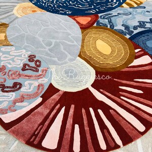 May include: A colorful, abstract area rug with a circular design. The rug features various shapes and patterns in shades of blue, brown, red, and gray. The design includes organic shapes and textured details, creating a modern and artistic look.
