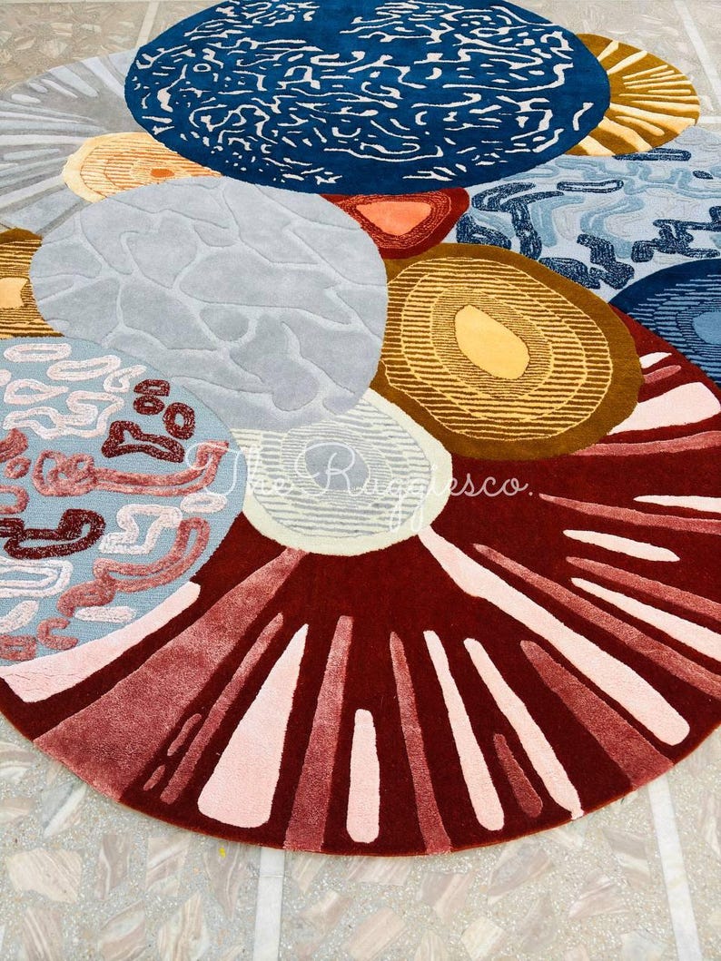 May include: A colorful, abstract area rug with a circular design. The rug features various shapes and patterns in shades of blue, brown, red, and gray. The design includes organic shapes and textured details, creating a modern and artistic look.