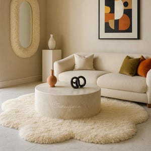 Op de afbeelding: Een woonkamer met een crèmekleurige bank, een ronde salontafel en een shaggy vloerkleed. De kamer bevat een ingelijst abstract kunstwerk, een grote ovale spiegel en decoratieve vazen. De salontafel heeft de tekst "Theruggiesco".