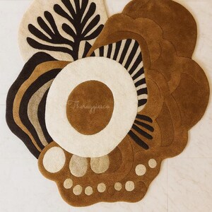 May include: An abstract, multi-colored rug with a floral design. The rug features a cream-colored background with brown, black, and tan accents. The rug has a unique shape and texture, with a variety of patterns and shapes.