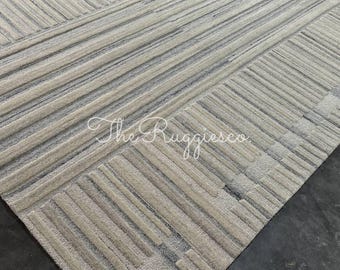 May include: A rectangular rug with a geometric pattern in beige and grey. The design features parallel and perpendicular lines creating a textured effect. The text "The Ruggiesco" is visible in the lower center of the rug.
